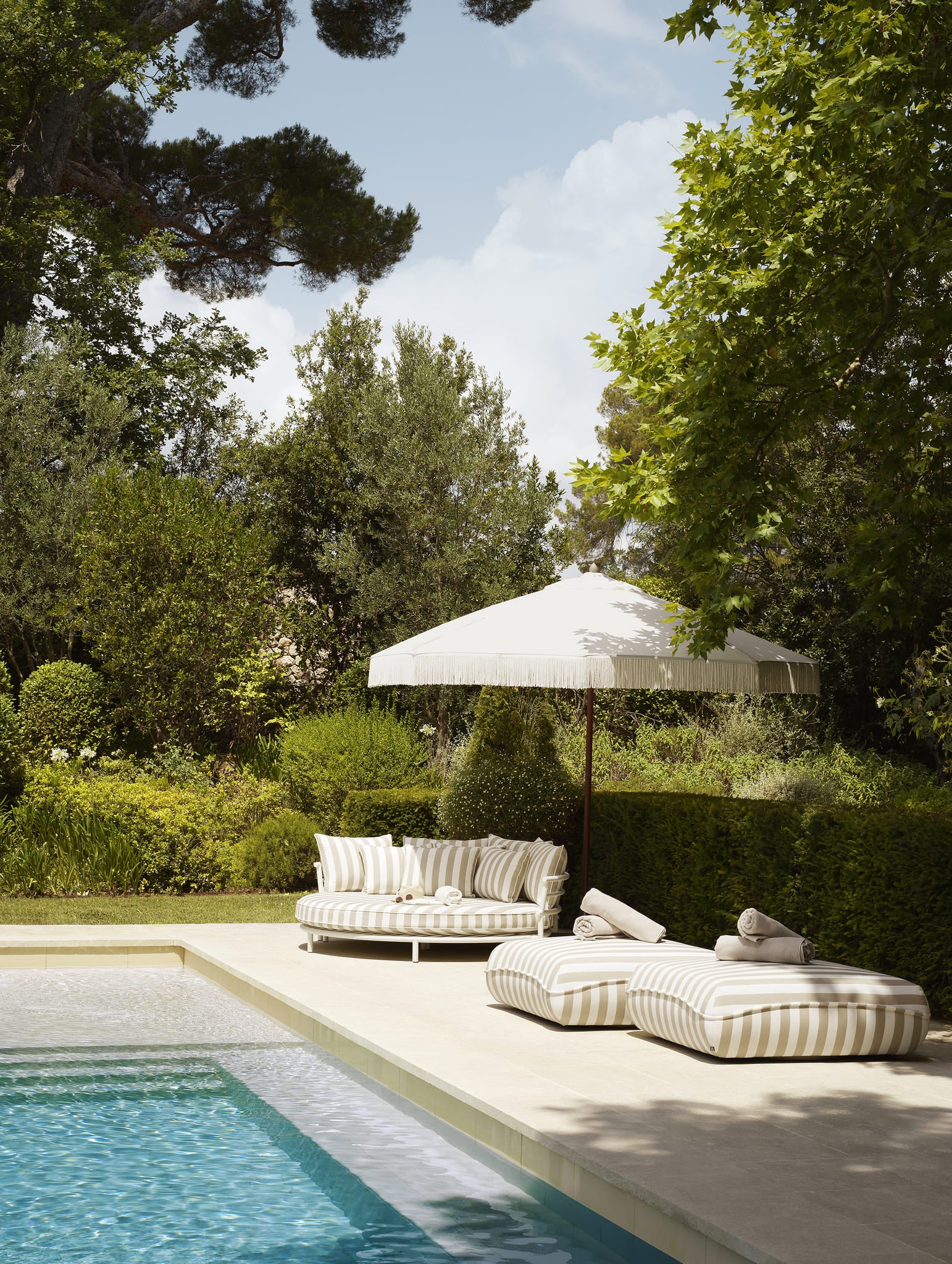 Exklusives Eichholtz Outdoor Daybed mit gestreiften Kissen und Sonnenschirm am Pool in grünem Gartenambiente.