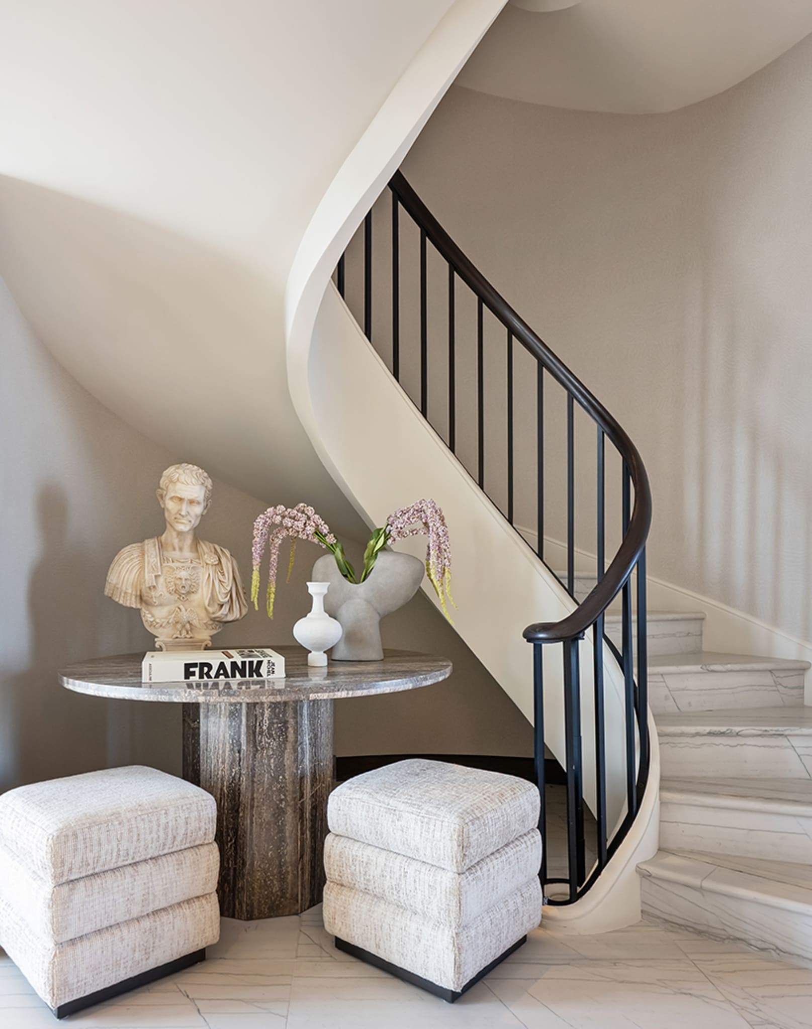 Elegant eingerichtetes Vorzimmer mit rundem Tisch, Skulptur und geschwungener Treppe.