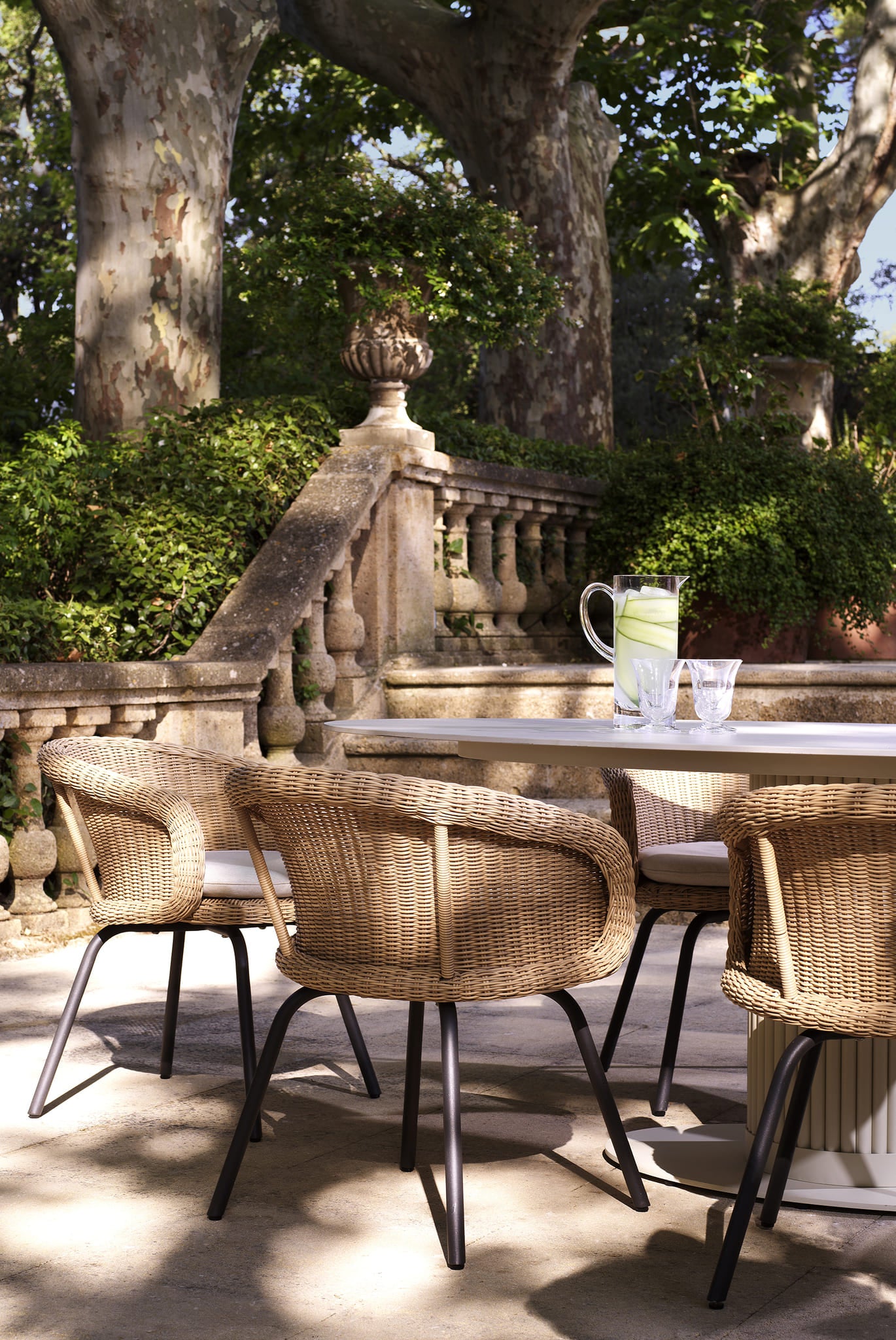 Stilvoller Eichholtz Gartentisch mit Rattan-Sesseln auf mediterraner Steinterrasse umgeben von üppiger Gartenlandschaft.