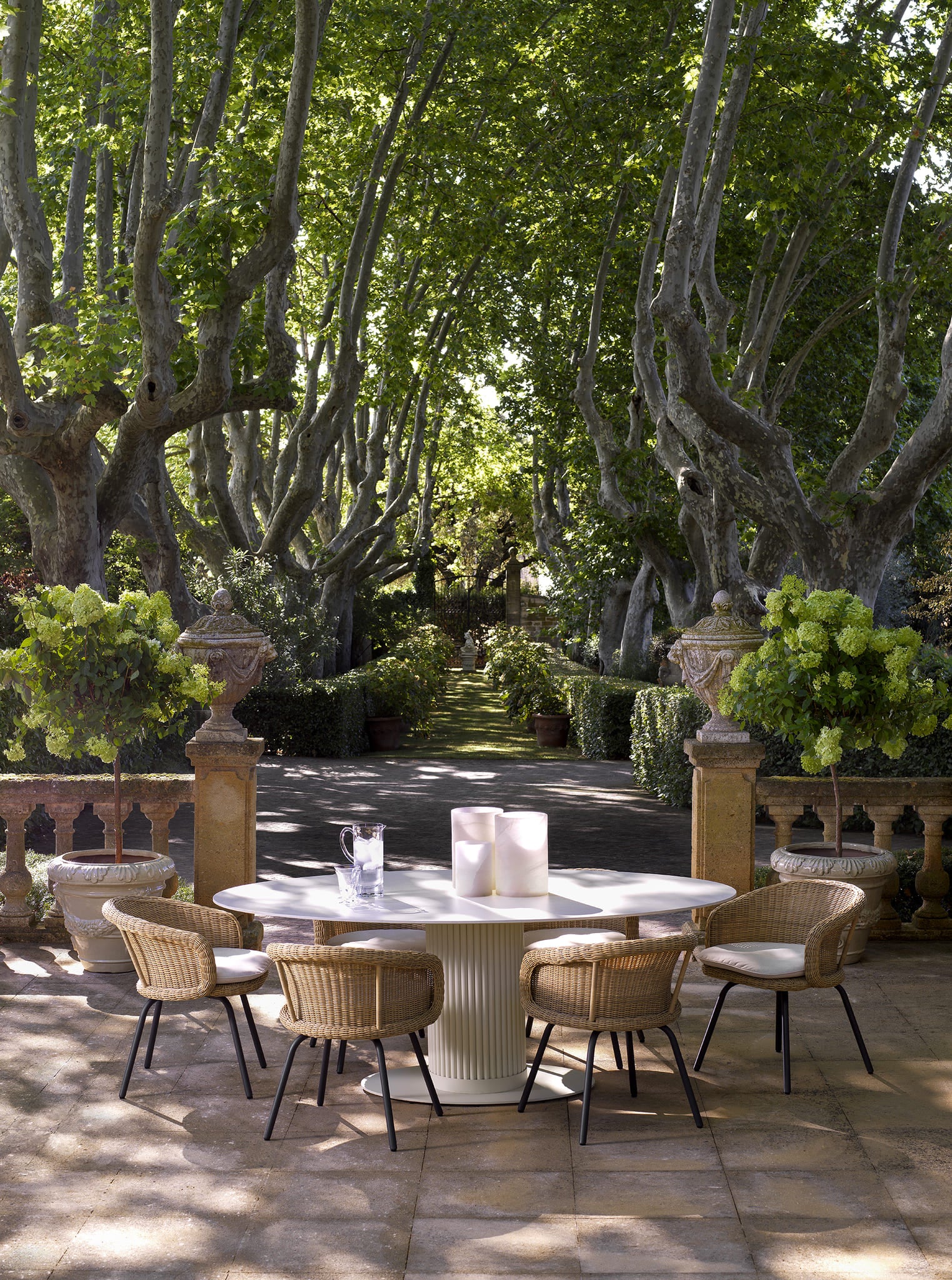 Eleganter Eichholtz Outdoor Esstisch mit Rattan-Stühlen auf einer sonnigen Gartenterrasse mit alten Platanen und klassischer Steinarchitektur.