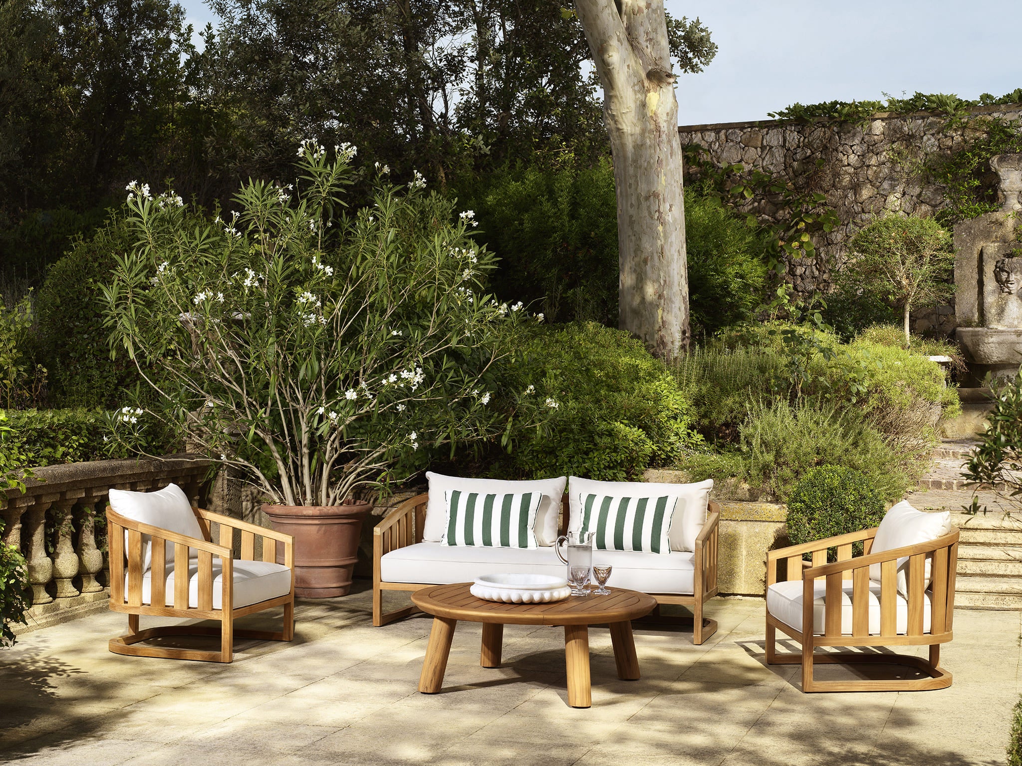 Luxuriöse Eichholtz Outdoor Lounge mit Holzsofa und Sesseln im klassischen Gartenambiente mit grünen Pflanzen und Natursteinmauer.