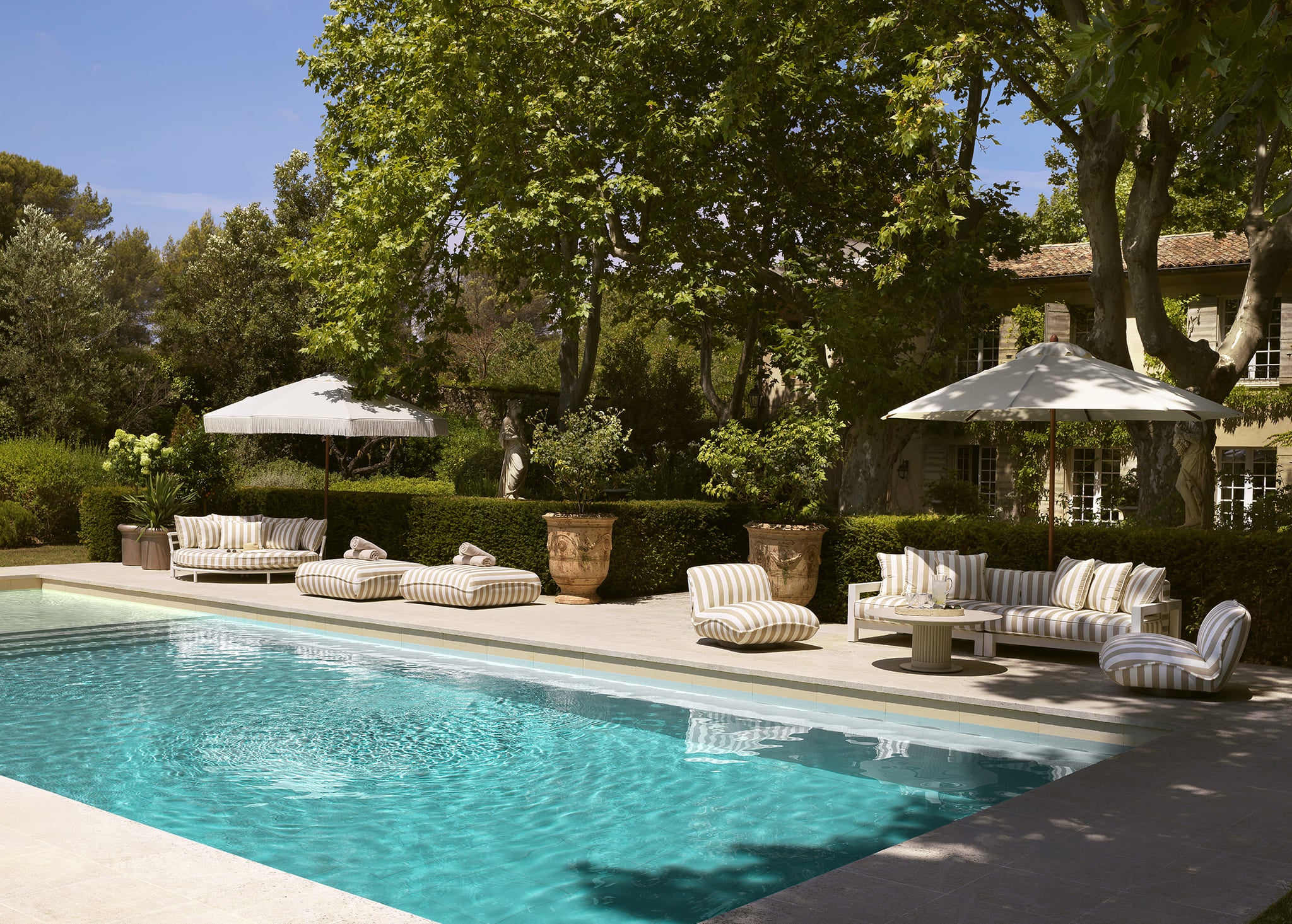 Stilvolle Eichholtz Outdoor Lounge mit gestreiften Polstern und Sonnenschirmen am Pool in mediterraner Gartenlandschaft.