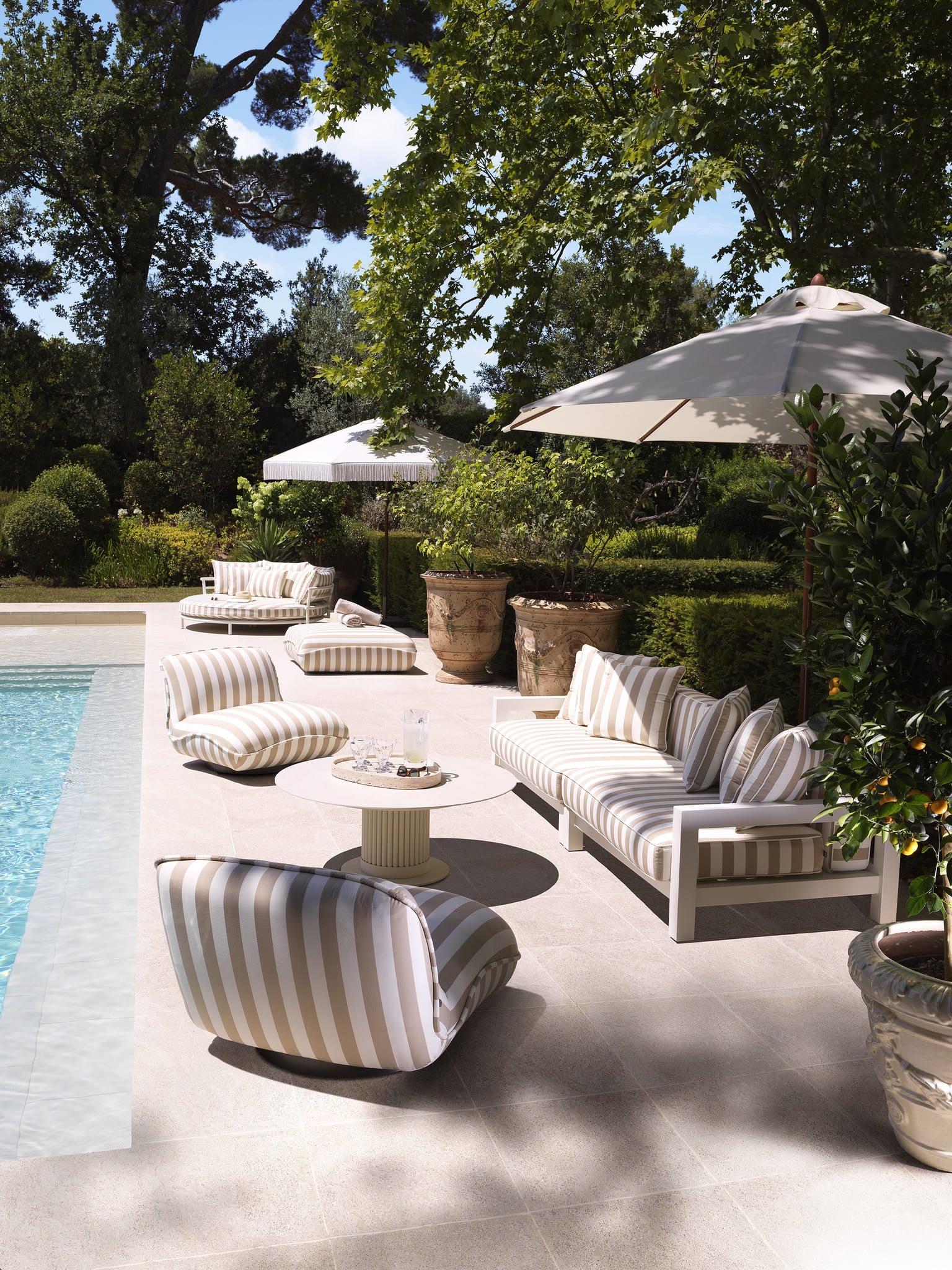 Exklusive Eichholtz Poollounge mit gestreiftem Outdoor-Sofa und Sesseln unter weißen Sonnenschirmen in sonniger Gartenkulisse.