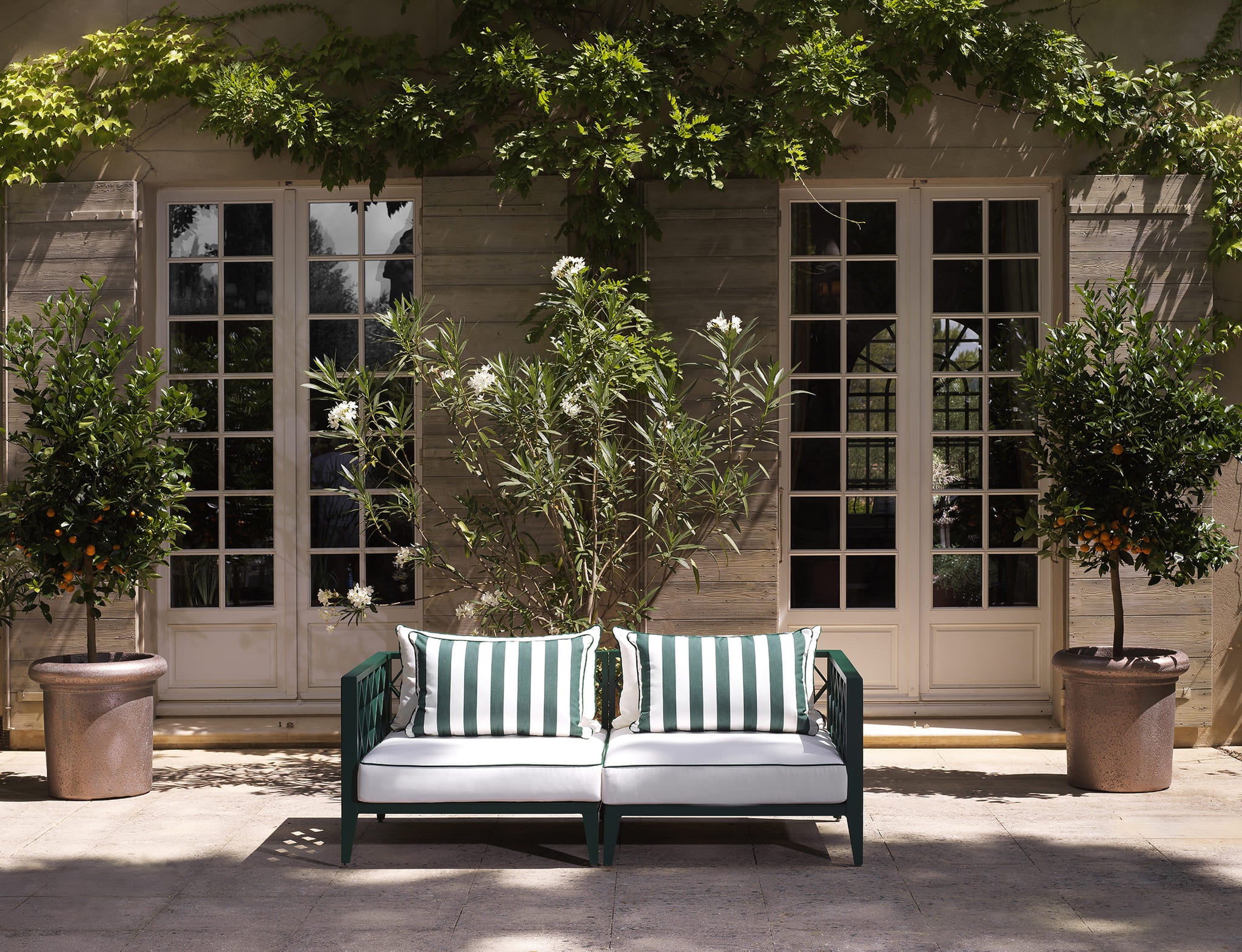 Zeitloses Eichholtz Gartensofa mit grün-weiß gestreiften Kissen vor klassischer Fassade mit französischen Türen und Orangenbäumen.
